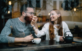 A man and a woman sit at a café table behind a window. The woman, smiling, holds the man's hand and brings it to her mouth as if kissing it. They are both enjoying beverages, with cups and a teapot on the table. The scene is cozy and warm.