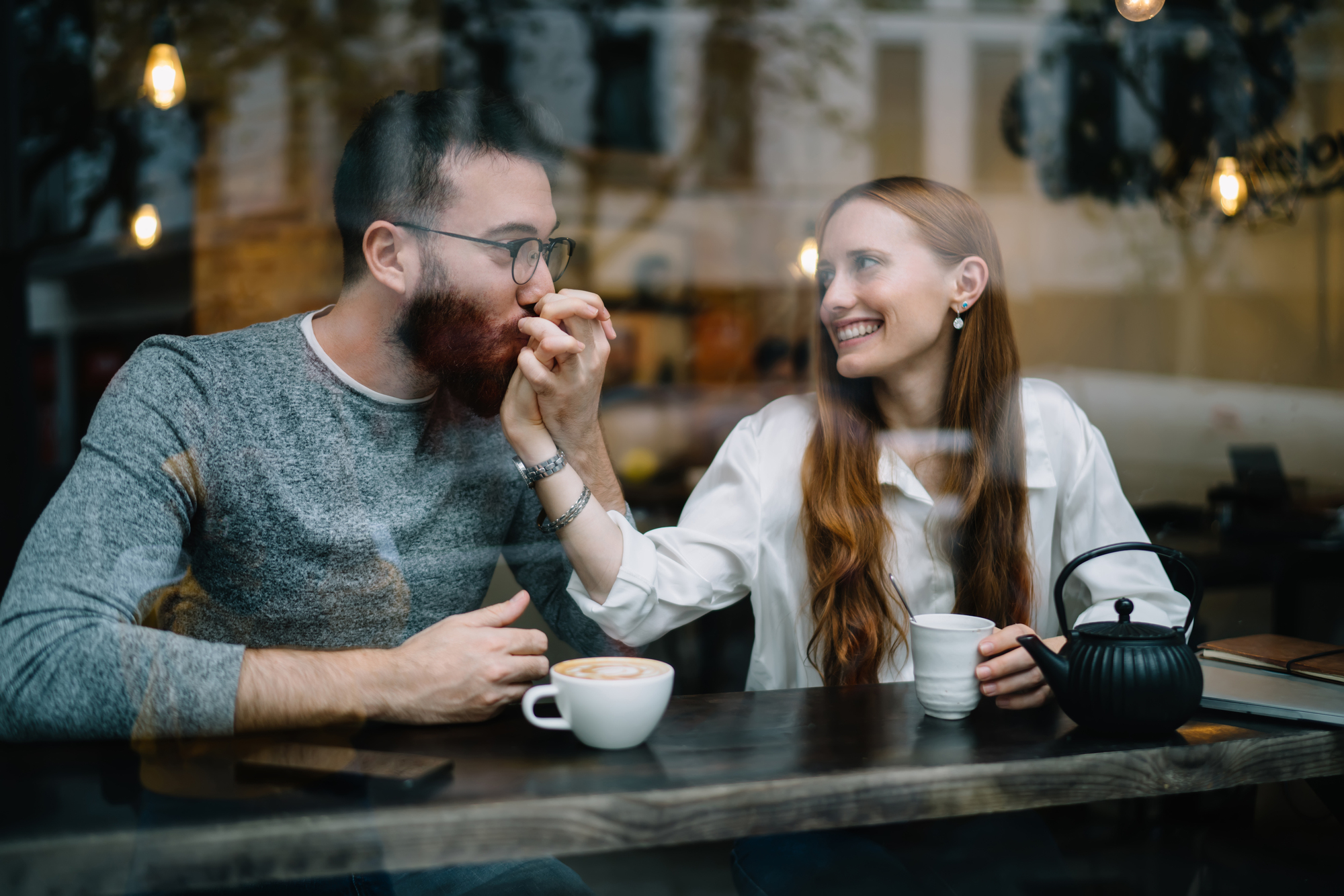 A man and a woman sit at a café table behind a window. The woman, smiling, holds the man's hand and brings it to her mouth as if kissing it. They are both enjoying beverages, with cups and a teapot on the table. The scene is cozy and warm.