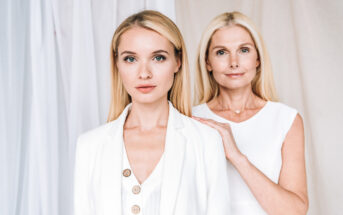 Two women with blonde hair stand together, both wearing white clothing. The younger woman in the foreground wears a blazer and looks into the camera with a neutral expression. The older woman stands behind her with one hand on her shoulder, also gazing at the camera.