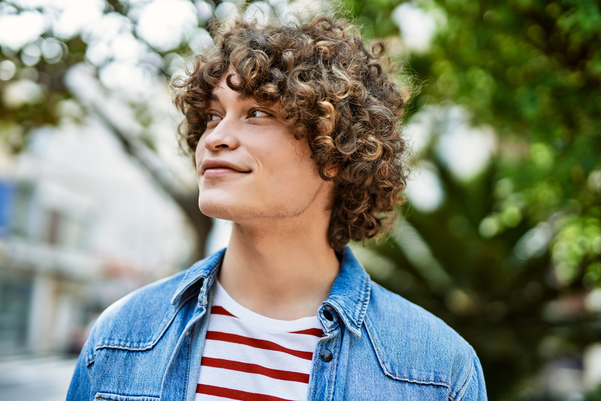 A person with curly hair, wearing a blue denim jacket over a red and white striped shirt, looks up and to the side while standing outdoors. The background is blurred, featuring green trees.