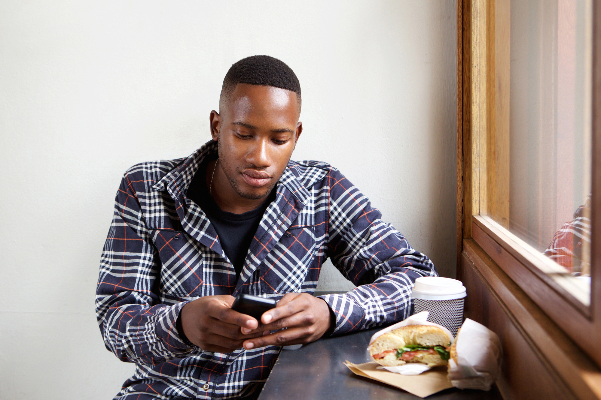 A person sits at a table near a window, looking at a smartphone. They are wearing a plaid shirt. On the table is a sandwich wrapped in paper and a coffee cup. The setting appears to be a casual dining spot.