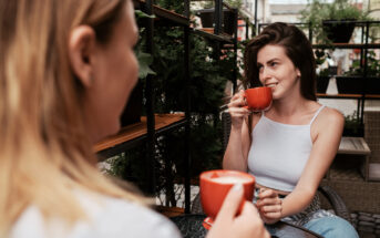 Two women are sitting at an outdoor café, each holding a red cup of coffee. One woman, with long brown hair, is smiling as she takes a sip. They are surrounded by greenery and potted plants, creating a relaxed and inviting atmosphere.