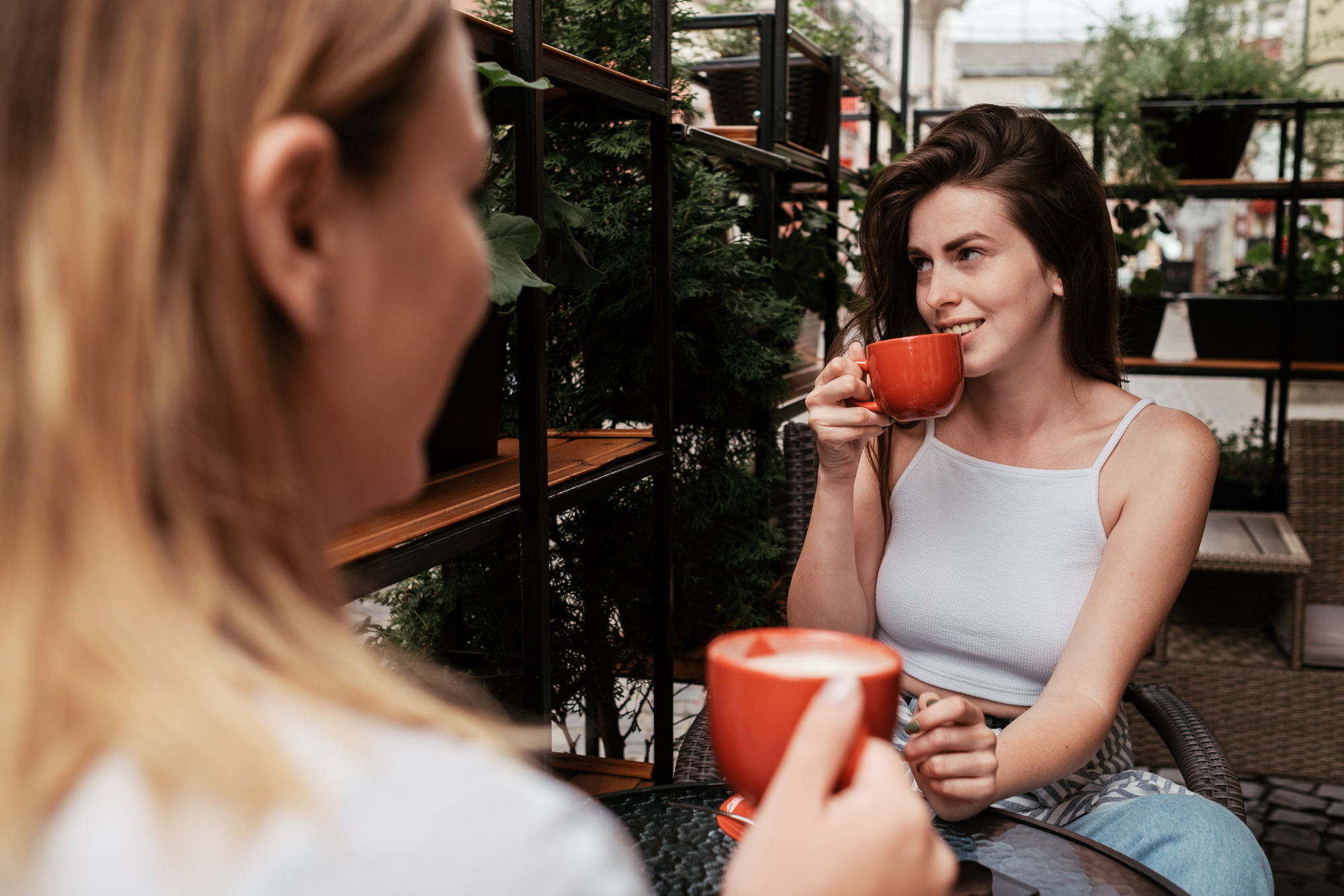 Two women are sitting at an outdoor café, each holding a red cup of coffee. One woman, with long brown hair, is smiling as she takes a sip. They are surrounded by greenery and potted plants, creating a relaxed and inviting atmosphere.