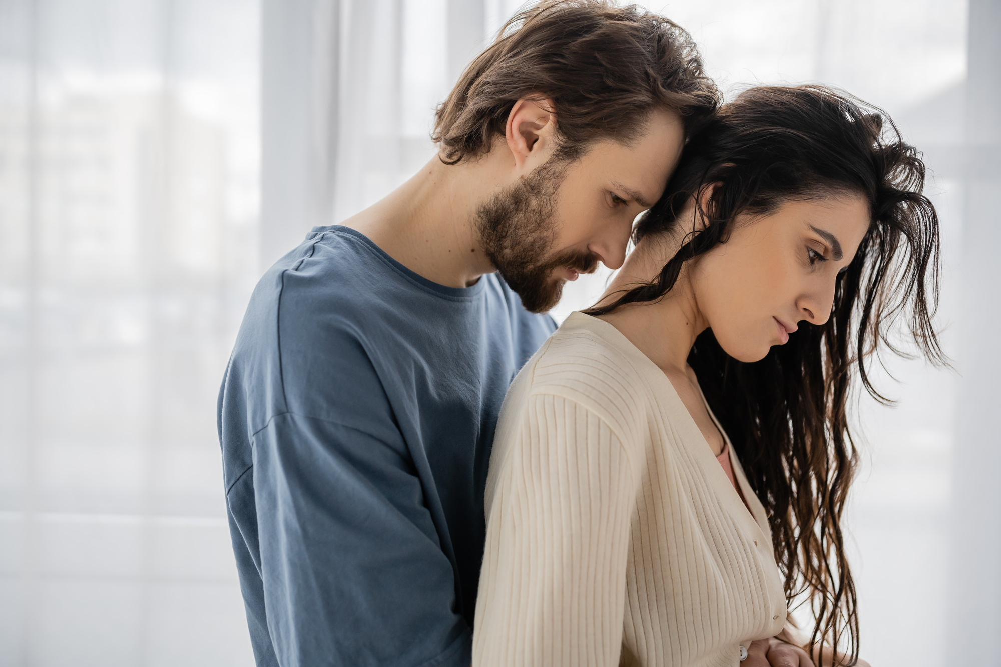 A man with a beard wearing a blue shirt embraces a woman with long, dark hair in a beige sweater. They both look pensive and are standing near a sheer curtain with a softly lit window in the background.