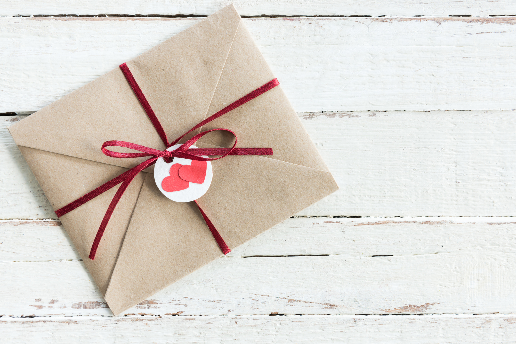 A brown kraft paper envelope tied with a red ribbon and a bow on a white wooden surface. A small white tag with two red hearts is attached to the ribbon.