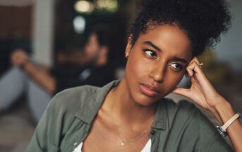 A woman with curly hair is seated indoors, resting her head on her hand, looking thoughtful or concerned. She wears a green blouse and a white top. In the background, a person is sitting on the floor, blurred and out of focus. The mood is contemplative.