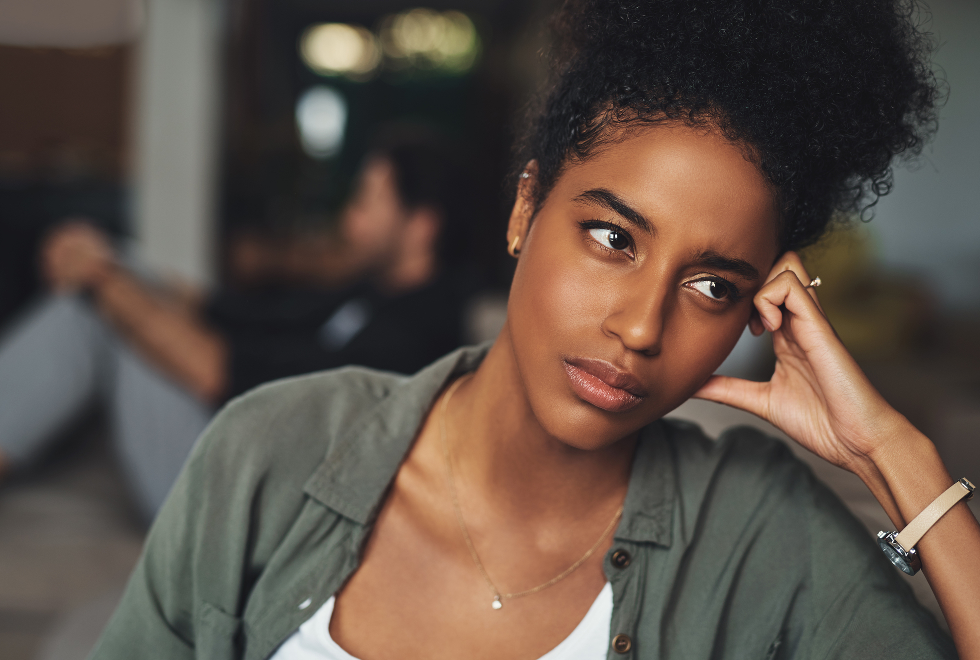 A woman with curly hair is seated indoors, resting her head on her hand, looking thoughtful or concerned. She wears a green blouse and a white top. In the background, a person is sitting on the floor, blurred and out of focus. The mood is contemplative.