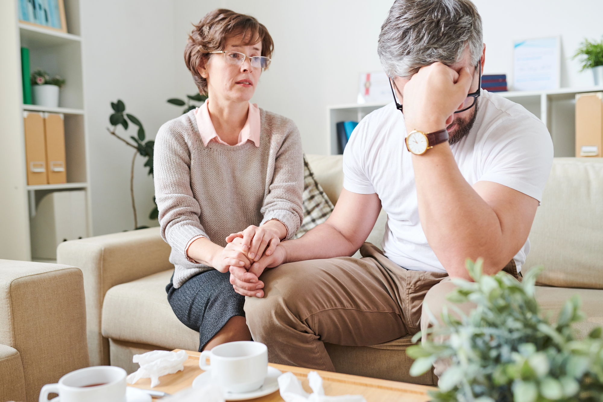 A woman with short hair and glasses sits on a beige couch, comforting a distressed man who is holding his head in his hand. They are surrounded by crumpled tissues, indicating emotional distress. A coffee table with mugs and papers is in front of them.