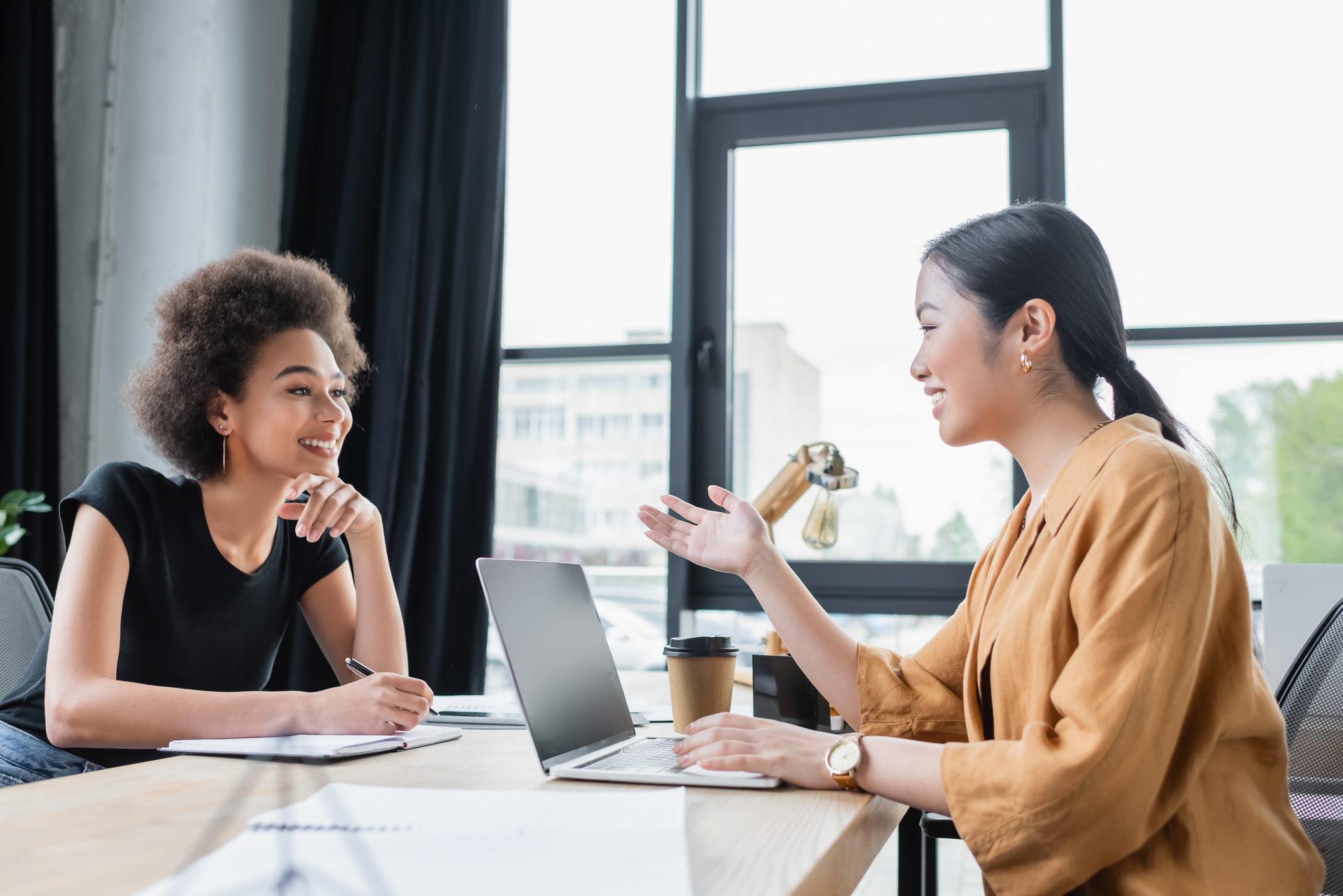 Two women are sitting at a wooden table in an office, engaged in a conversation. One woman is writing in a notebook with a pen while the other is speaking and gesturing with her hand. They both have laptops open in front of them and a cup of coffee nearby.