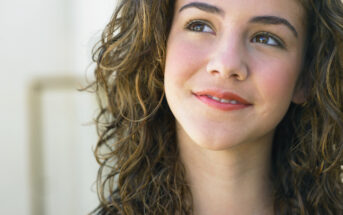 A young woman with curly brown hair and rosy cheeks is looking slightly upwards and smiling softly. She is wearing a blue top, and her expression conveys a sense of contentment or contemplation. The background is a neutral, light color, slightly blurred.