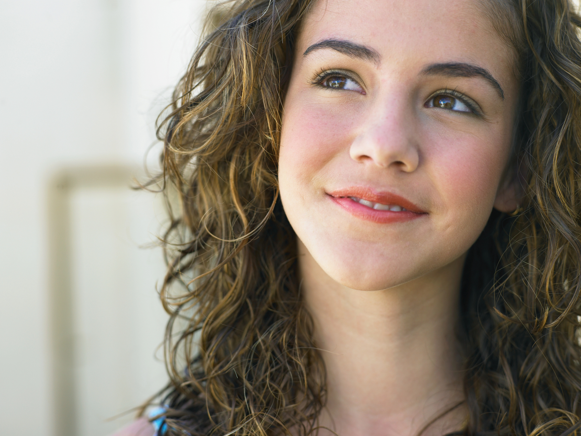 A young woman with curly brown hair and rosy cheeks is looking slightly upwards and smiling softly. She is wearing a blue top, and her expression conveys a sense of contentment or contemplation. The background is a neutral, light color, slightly blurred.