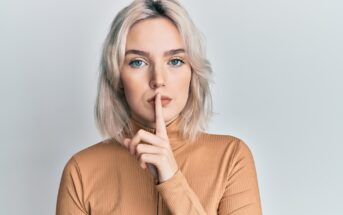 A person with shoulder-length blonde hair and blue eyes is standing against a plain background, with their index finger pressed to their lips in a "shushing" gesture. They are wearing a light brown turtleneck.