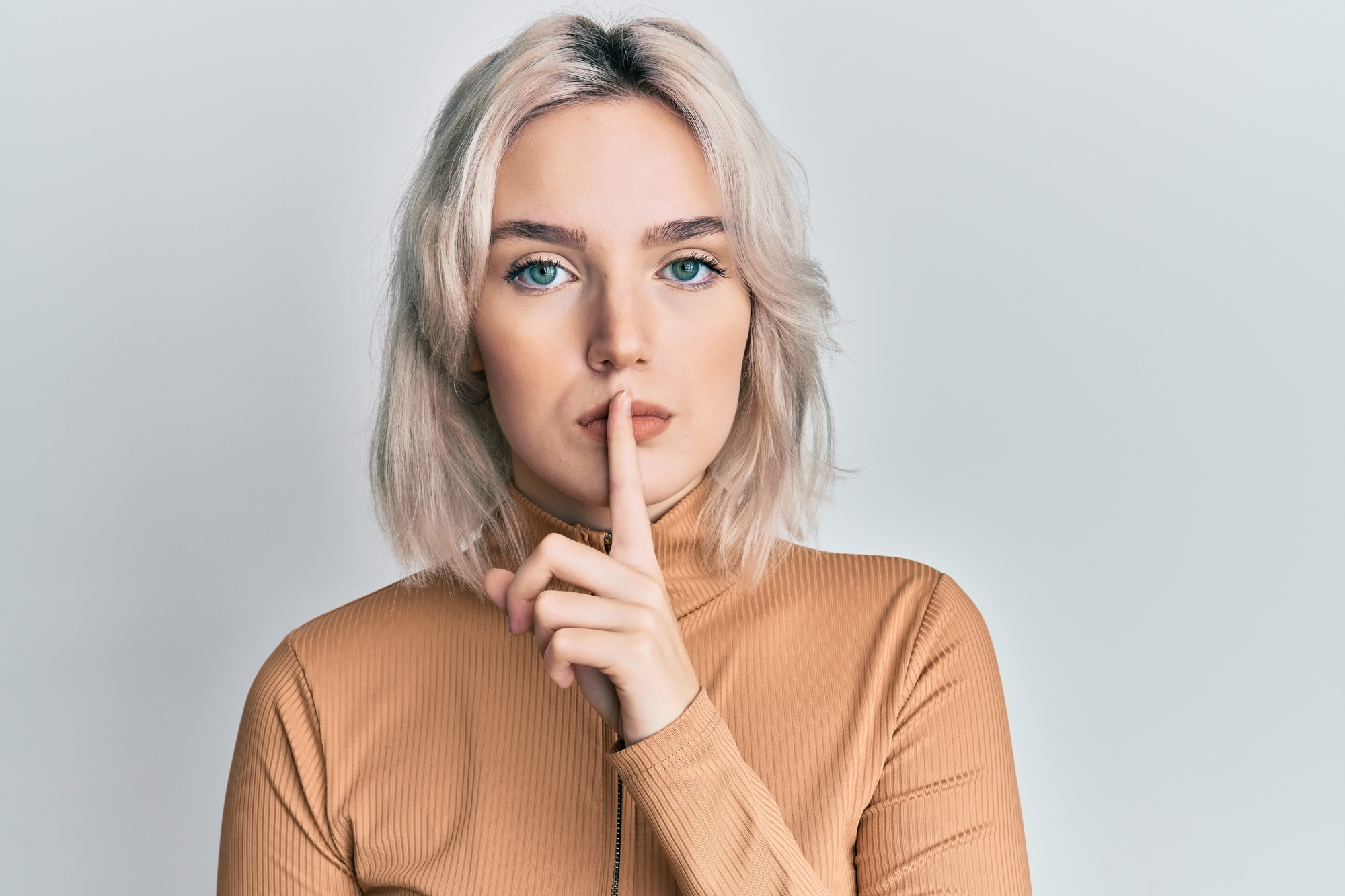 A person with shoulder-length blonde hair and blue eyes is standing against a plain background, with their index finger pressed to their lips in a "shushing" gesture. They are wearing a light brown turtleneck.