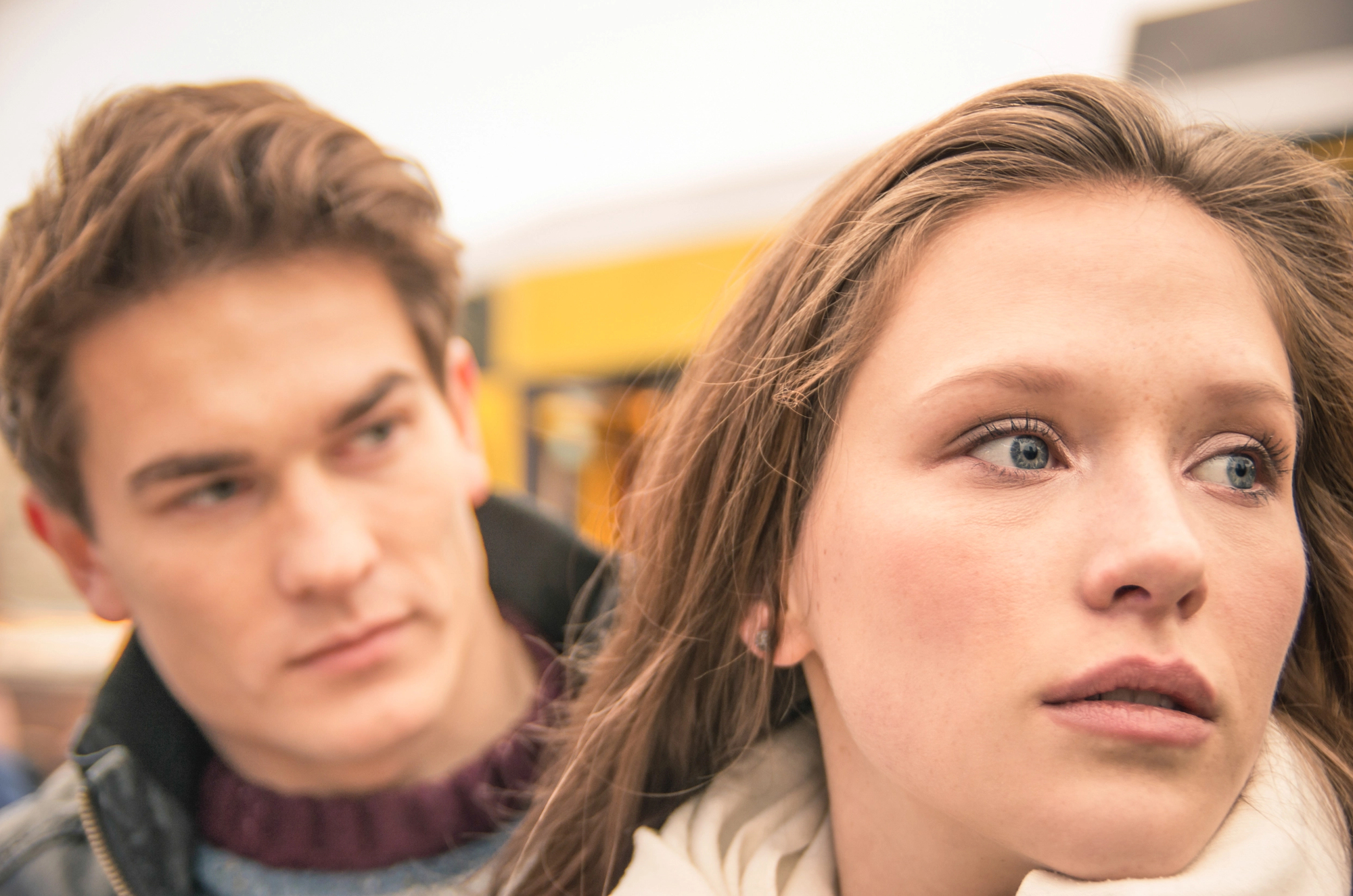 A young woman with long light brown hair looks pensively into the distance. A young man with short brown hair stands closely behind her, looking at her with a concerned expression. The background is slightly blurred, featuring a yellow structure.