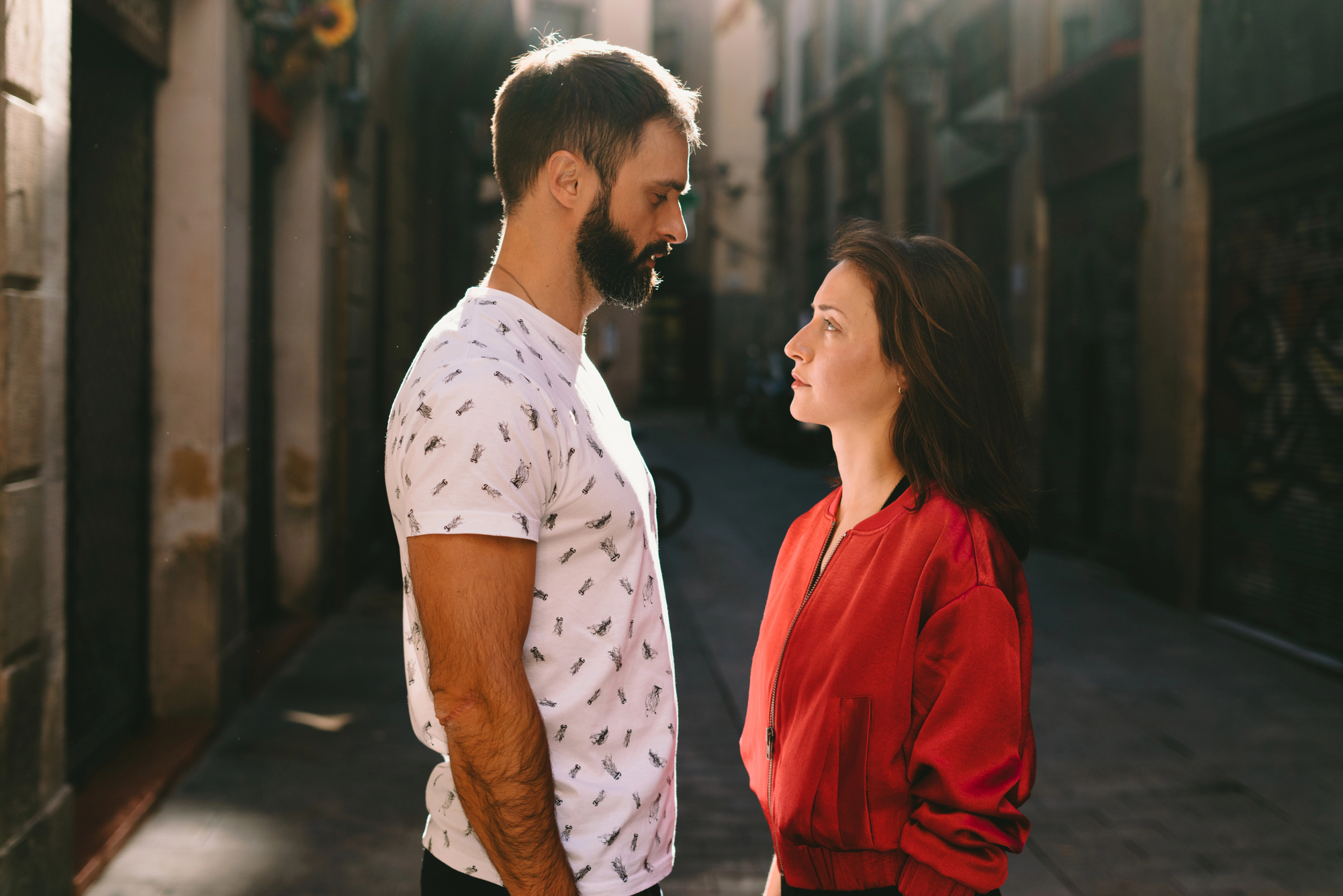 A man with a beard, wearing a white patterned t-shirt, faces a woman with long hair in a red jacket. They stand close to each other, looking into each other's eyes, on a narrow, sunlit street lined with old buildings.