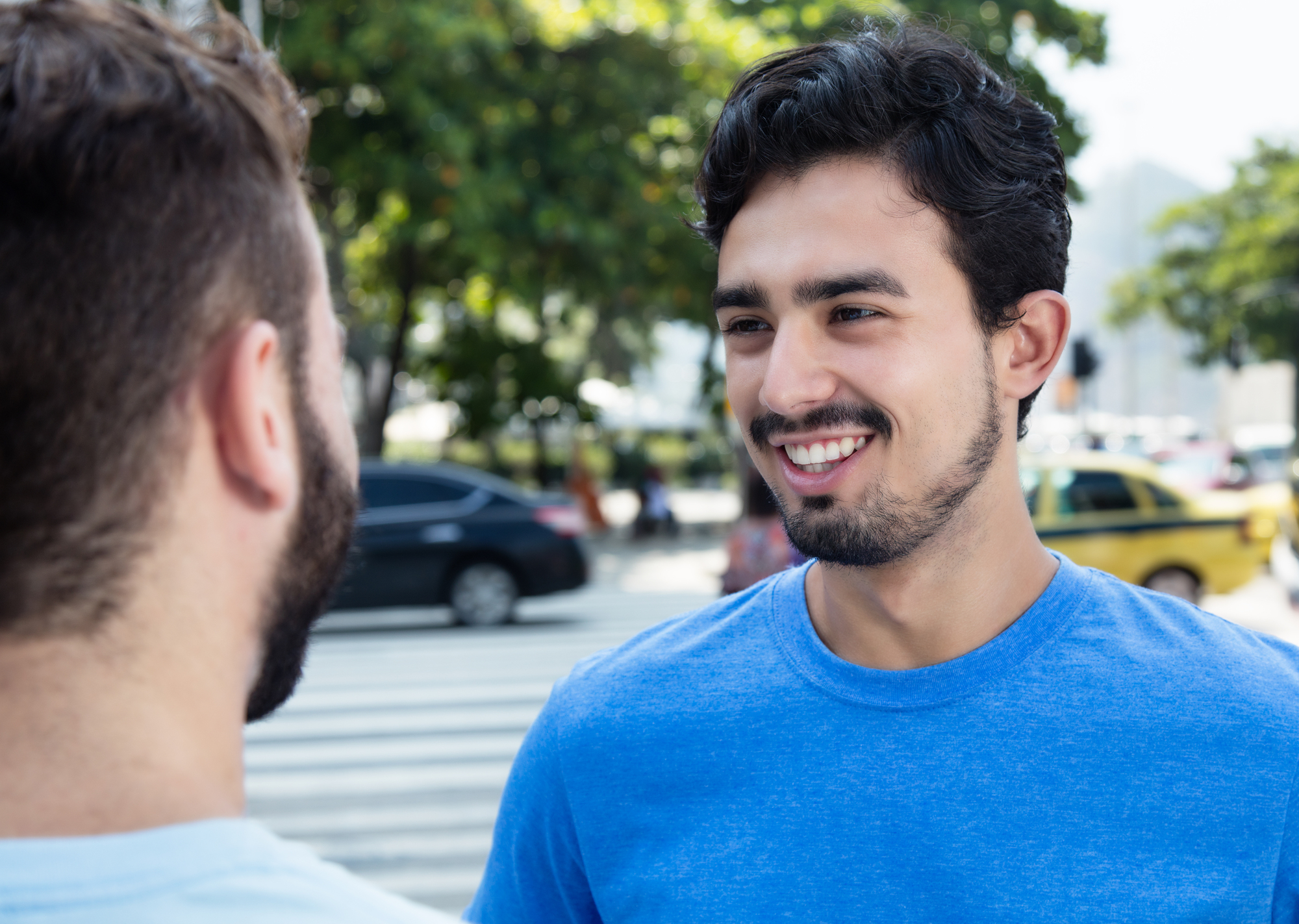 Two men are talking on a street. The man facing the camera is smiling and has dark hair with a short beard, wearing a blue t-shirt. The other man's back is to the camera, and he has short brown hair and a beard. Cars and trees are visible in the background.