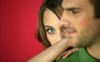 A close-up of a woman with brown hair looking directly at the camera with a neutral expression, her arm resting on the shoulder of a man in the foreground who is wearing a green shirt. The background is a solid red color.