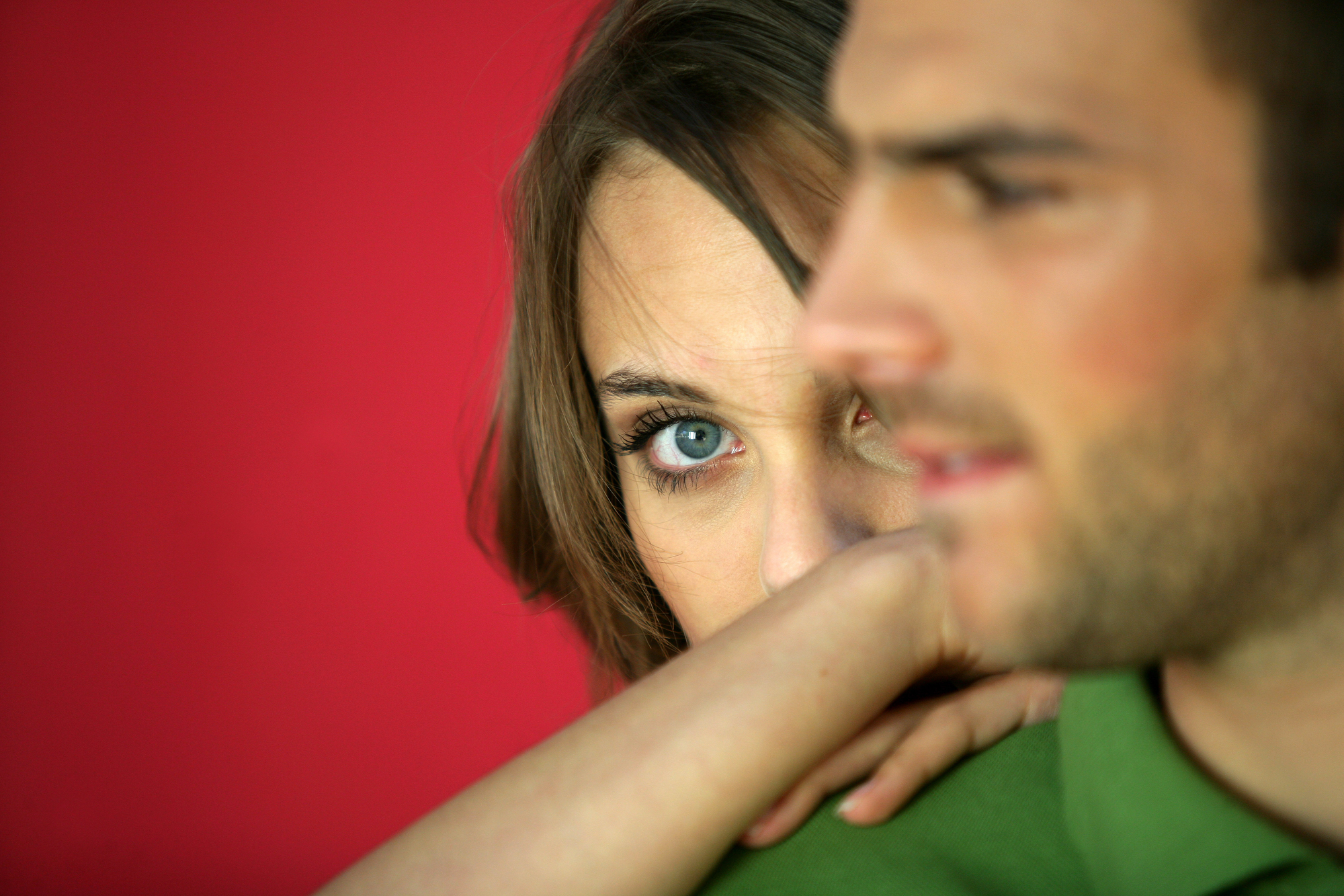 A close-up of a woman with brown hair looking directly at the camera with a neutral expression, her arm resting on the shoulder of a man in the foreground who is wearing a green shirt. The background is a solid red color.