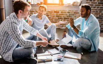 A group of four young adults, three males and one female, sit on the floor of a room with brick walls and large windows. They are engaged in discussion, with notebooks, pens, and smartphones spread out around them. Sunlight streams in, creating a warm atmosphere.