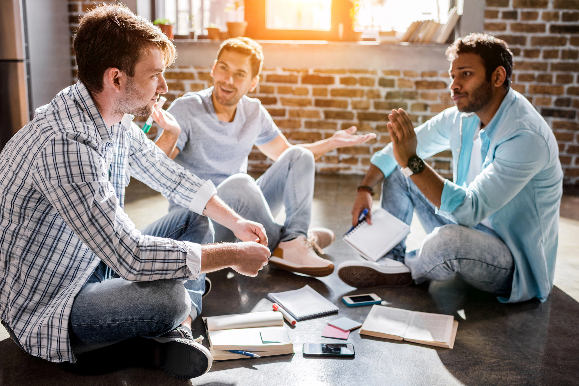 A group of four young adults, three males and one female, sit on the floor of a room with brick walls and large windows. They are engaged in discussion, with notebooks, pens, and smartphones spread out around them. Sunlight streams in, creating a warm atmosphere.