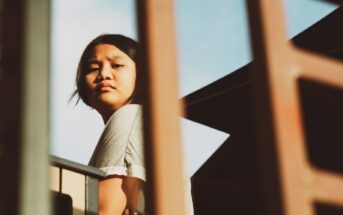 A person with long, dark hair wearing a grey t-shirt looks pensively to the side while standing on a balcony, with architectural elements and clear sky in the background. The image is framed by foreground structure, adding depth and focus on the subject.