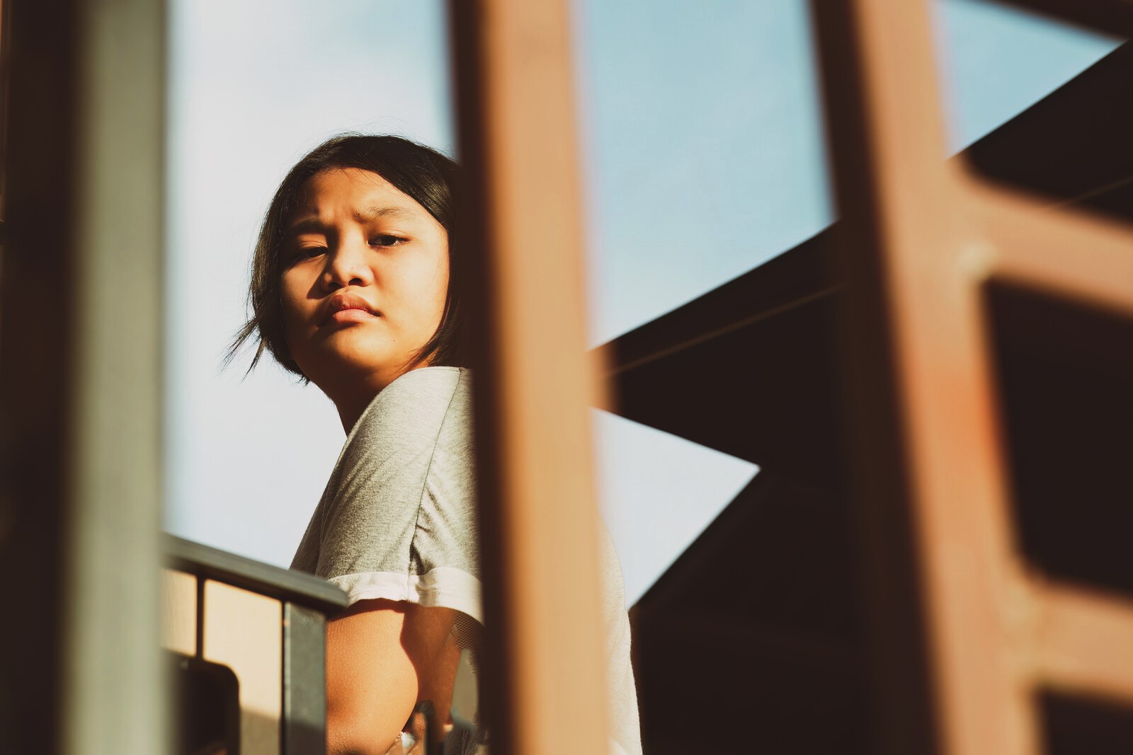 A person with long, dark hair wearing a grey t-shirt looks pensively to the side while standing on a balcony, with architectural elements and clear sky in the background. The image is framed by foreground structure, adding depth and focus on the subject.