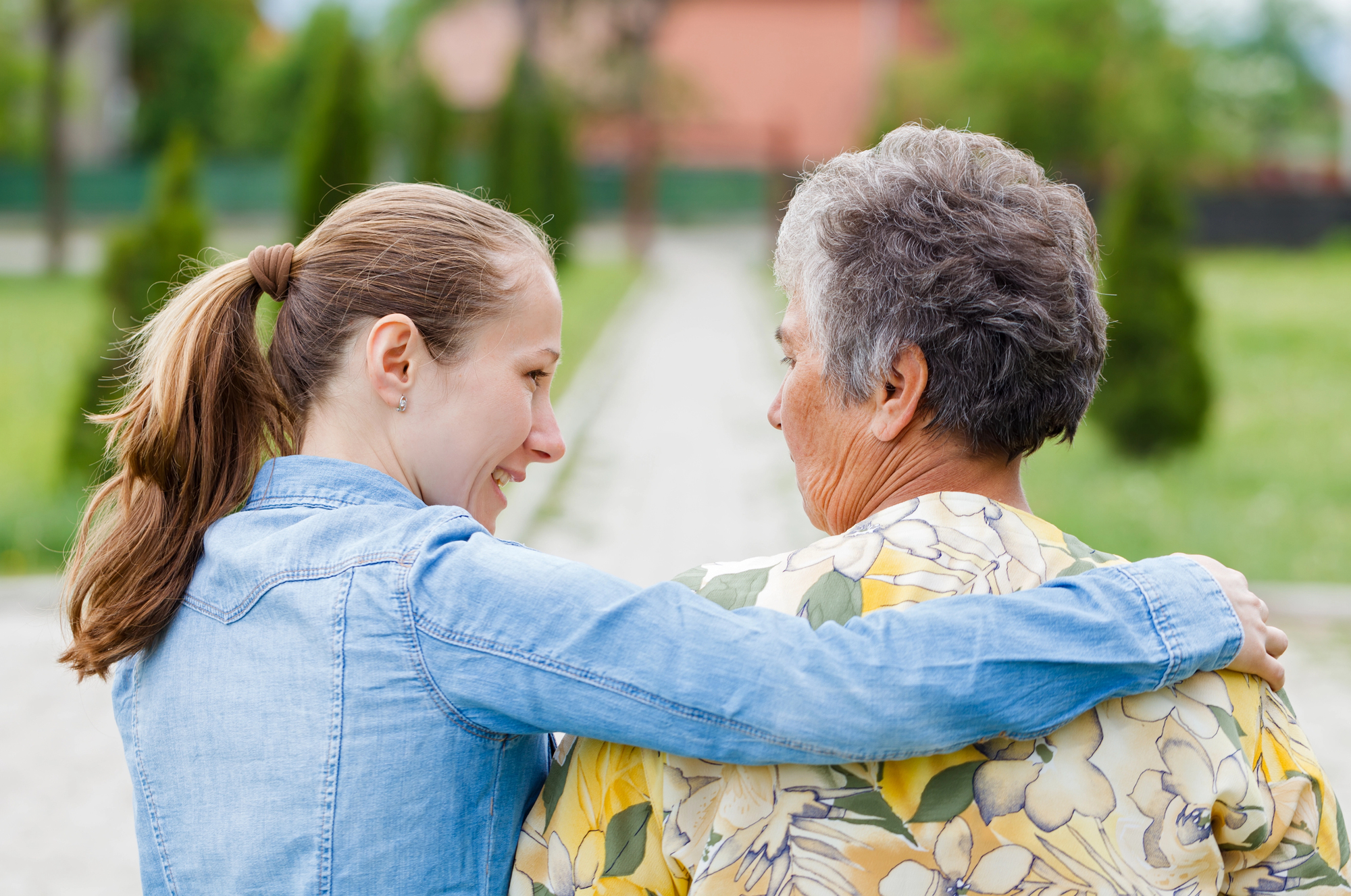 A young woman with a ponytail and a denim jacket has her arm around an older woman with short gray hair and a floral blouse. They are outdoors on a pathway lined with greenery, smiling and looking at each other. The background is softly blurred.