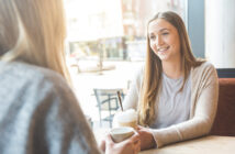 Two people are sitting at a table in a cafe, engaging in conversation. The person facing the camera is smiling and holding a drink topped with whipped cream, while the other person has their back turned, also holding a cup. The background shows large windows and street view.