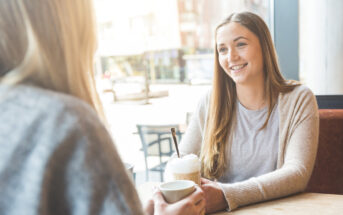 Two people are sitting at a table in a cafe, engaging in conversation. The person facing the camera is smiling and holding a drink topped with whipped cream, while the other person has their back turned, also holding a cup. The background shows large windows and street view.