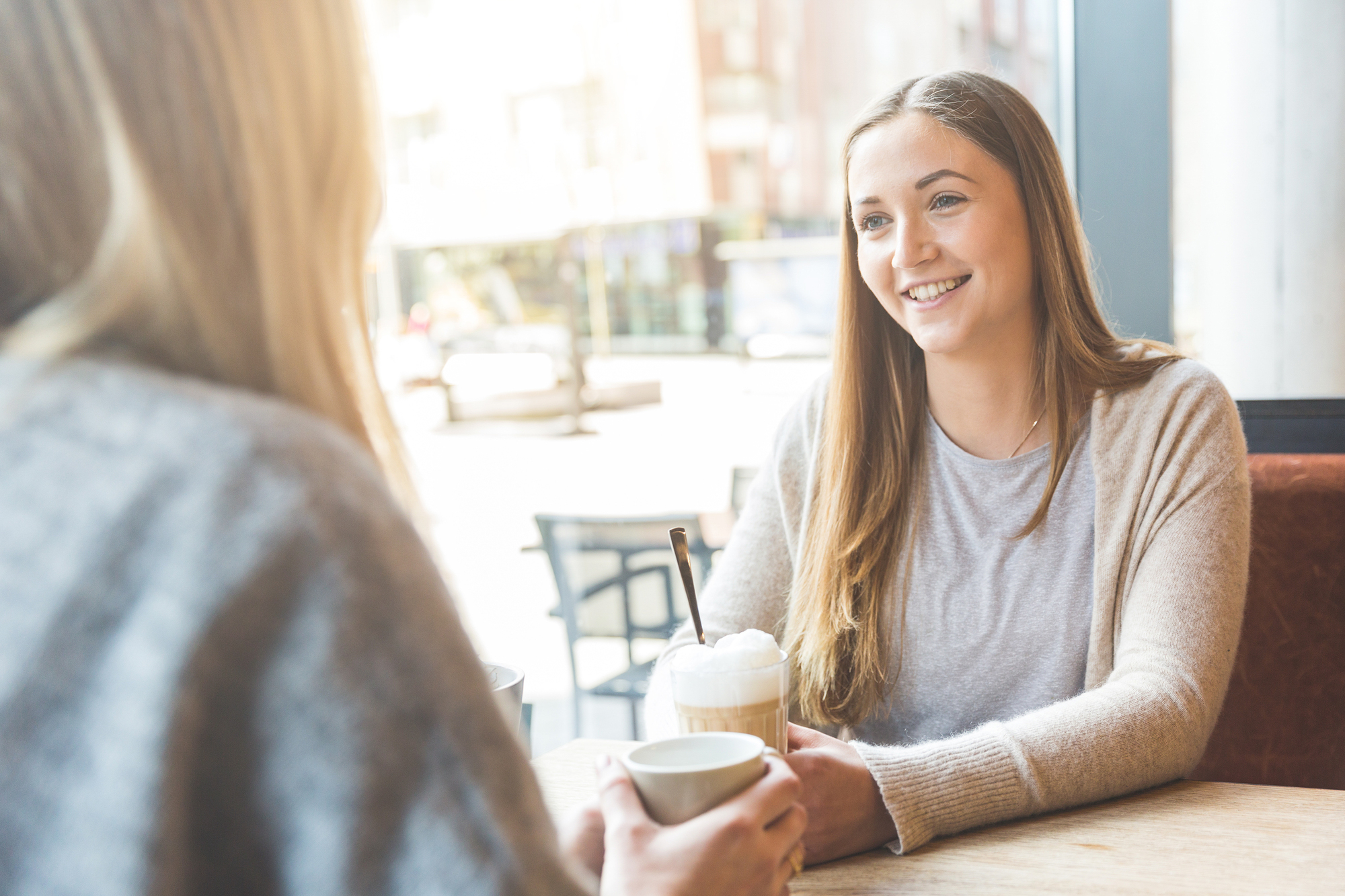 Two people are sitting at a table in a cafe, engaging in conversation. The person facing the camera is smiling and holding a drink topped with whipped cream, while the other person has their back turned, also holding a cup. The background shows large windows and street view.