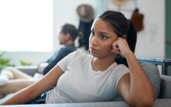 A young woman with long dark hair, wearing a light gray t-shirt, sits on a couch looking upset, resting her head on her hand. In the blurred background, a man sits on another couch, facing away. They appear to be in a tense or reflective moment.