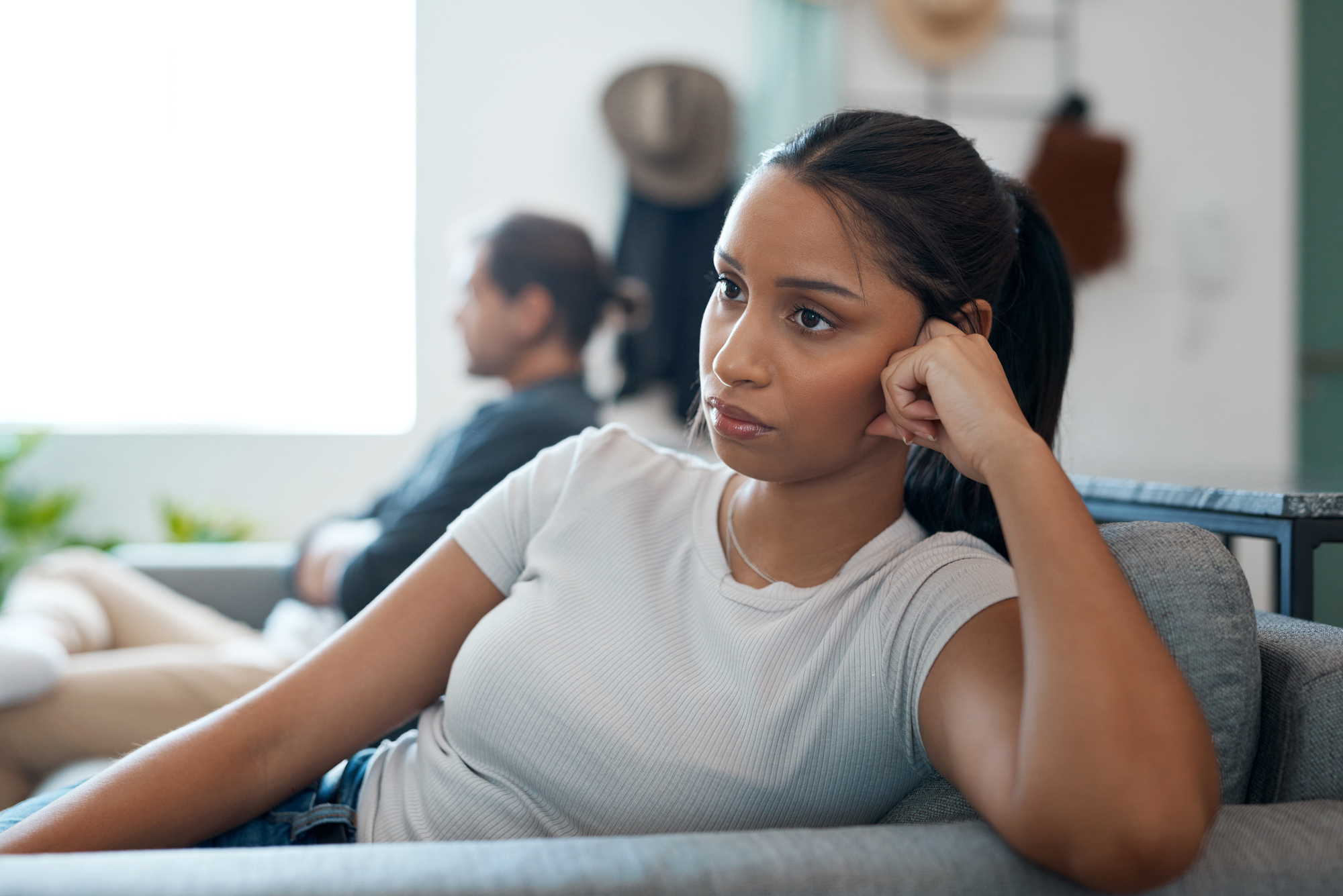 A young woman with long dark hair, wearing a light gray t-shirt, sits on a couch looking upset, resting her head on her hand. In the blurred background, a man sits on another couch, facing away. They appear to be in a tense or reflective moment.