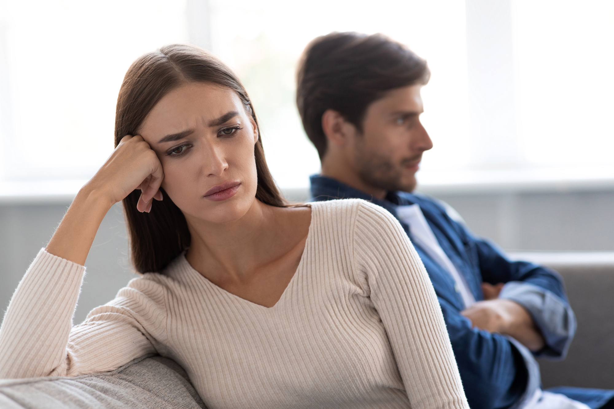 A woman with long brown hair wearing a white sweater sits on a couch, resting her head on her left hand and looking upset. In the background, a man with short brown hair in a blue shirt sits with his arms crossed, looking away. Both appear to be in distress.