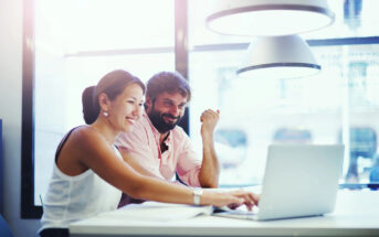 A woman and a man work together at a desk, smiling and looking at a laptop. The woman is typing while the man observes. They are in a bright, modern office with large windows in the background and hanging lamps above them.