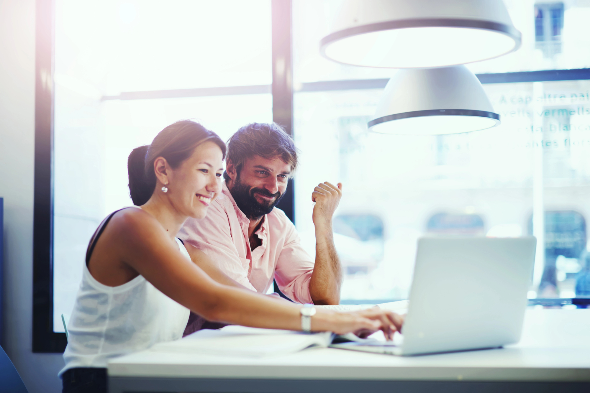 A woman and a man work together at a desk, smiling and looking at a laptop. The woman is typing while the man observes. They are in a bright, modern office with large windows in the background and hanging lamps above them.