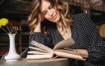 A woman with wavy brown hair and a black dress with white polka dots is sitting at a table, reading a book. She is leaning her head on her hand and smiling softly. There is a small vase with a yellow flower on the table beside her.