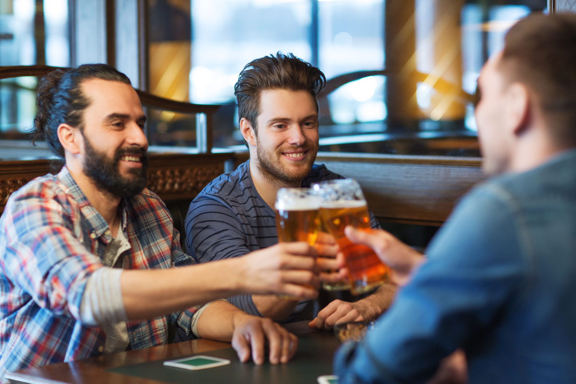 Three men are sitting at a bar, raising their glasses for a cheers. They are dressed casually and appear to be enjoying their time together. There is a wooden bar counter and some background patrons blurred out.