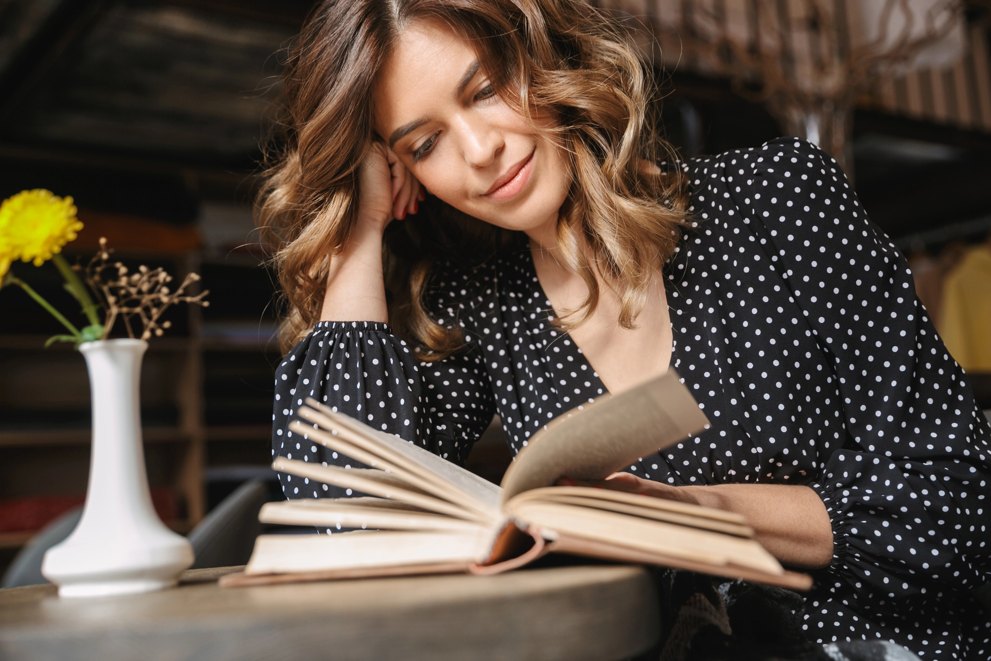 A woman with wavy brown hair and a black dress with white polka dots is sitting at a table, reading a book. She is leaning her head on her hand and smiling softly. There is a small vase with a yellow flower on the table beside her.