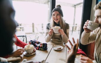 Three people are seated around a table in a cozy cafe, enjoying hot drinks. A woman in a knit hat and sweater is smiling while holding a mug. The background shows large windows with a slightly blurred outdoor view. Knitted scarves and gloves are on the table.