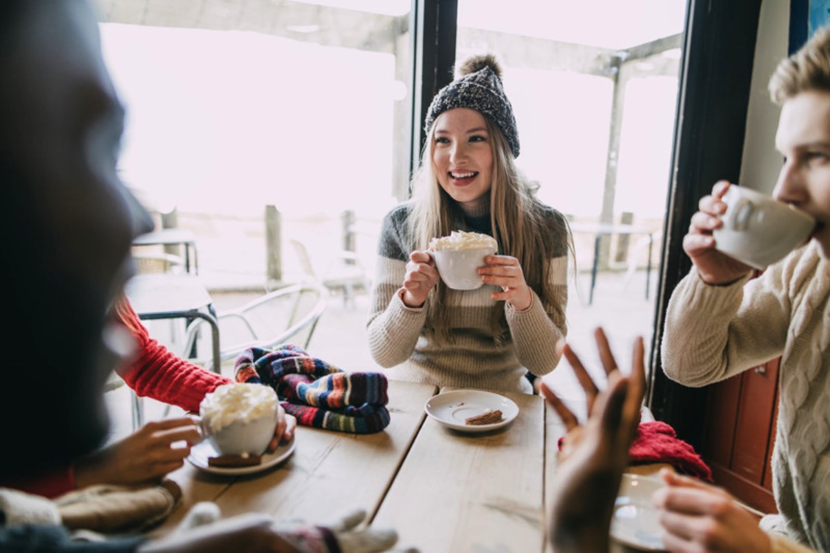 Three people are seated around a table in a cozy cafe, enjoying hot drinks. A woman in a knit hat and sweater is smiling while holding a mug. The background shows large windows with a slightly blurred outdoor view. Knitted scarves and gloves are on the table.