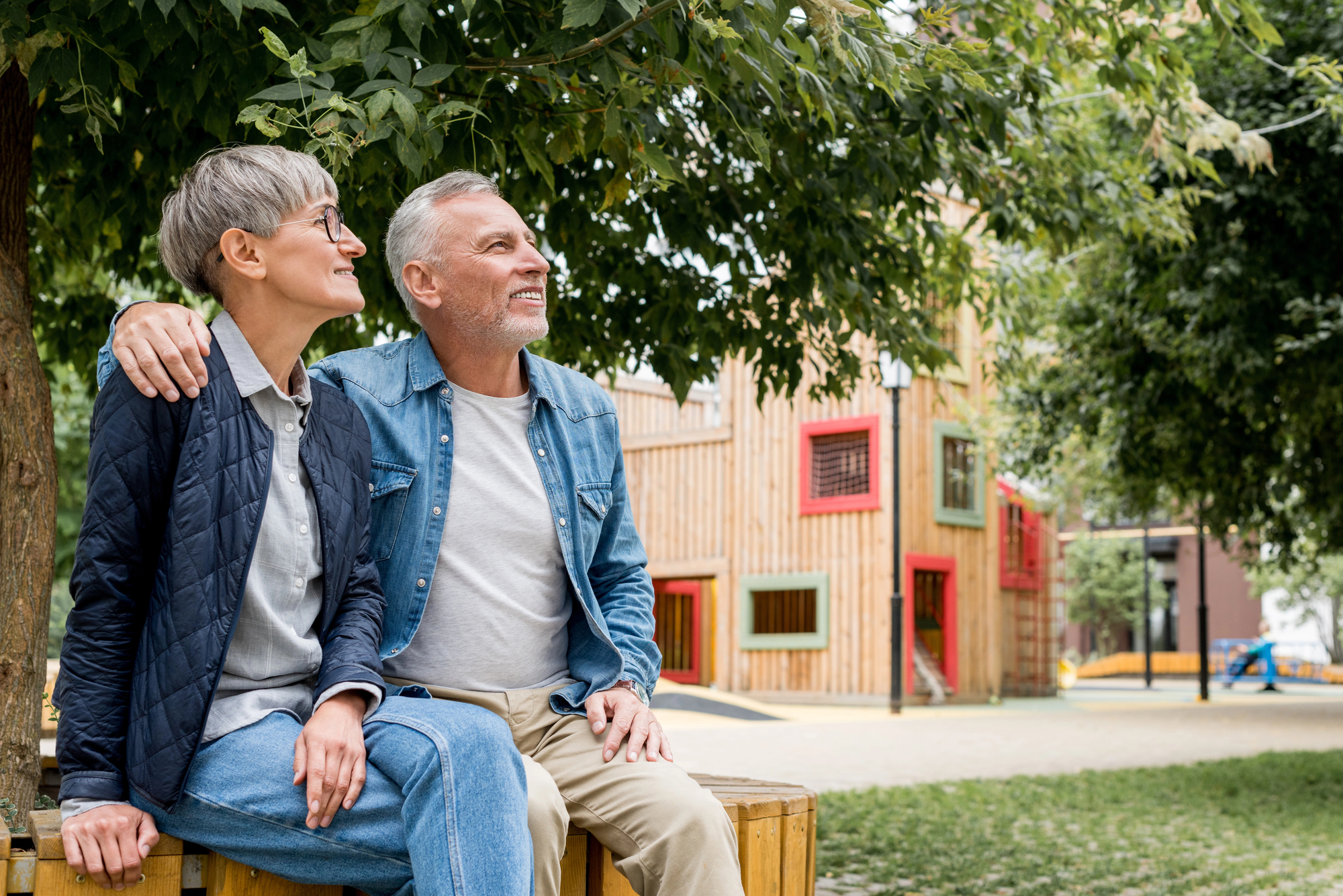 An elderly couple sits on a bench under a tree, looking up and smiling. Both are casually dressed; the woman wears a dark jacket and jeans, while the man has on a blue denim shirt and khaki pants. Behind them, there's a colorful wooden building and greenery.