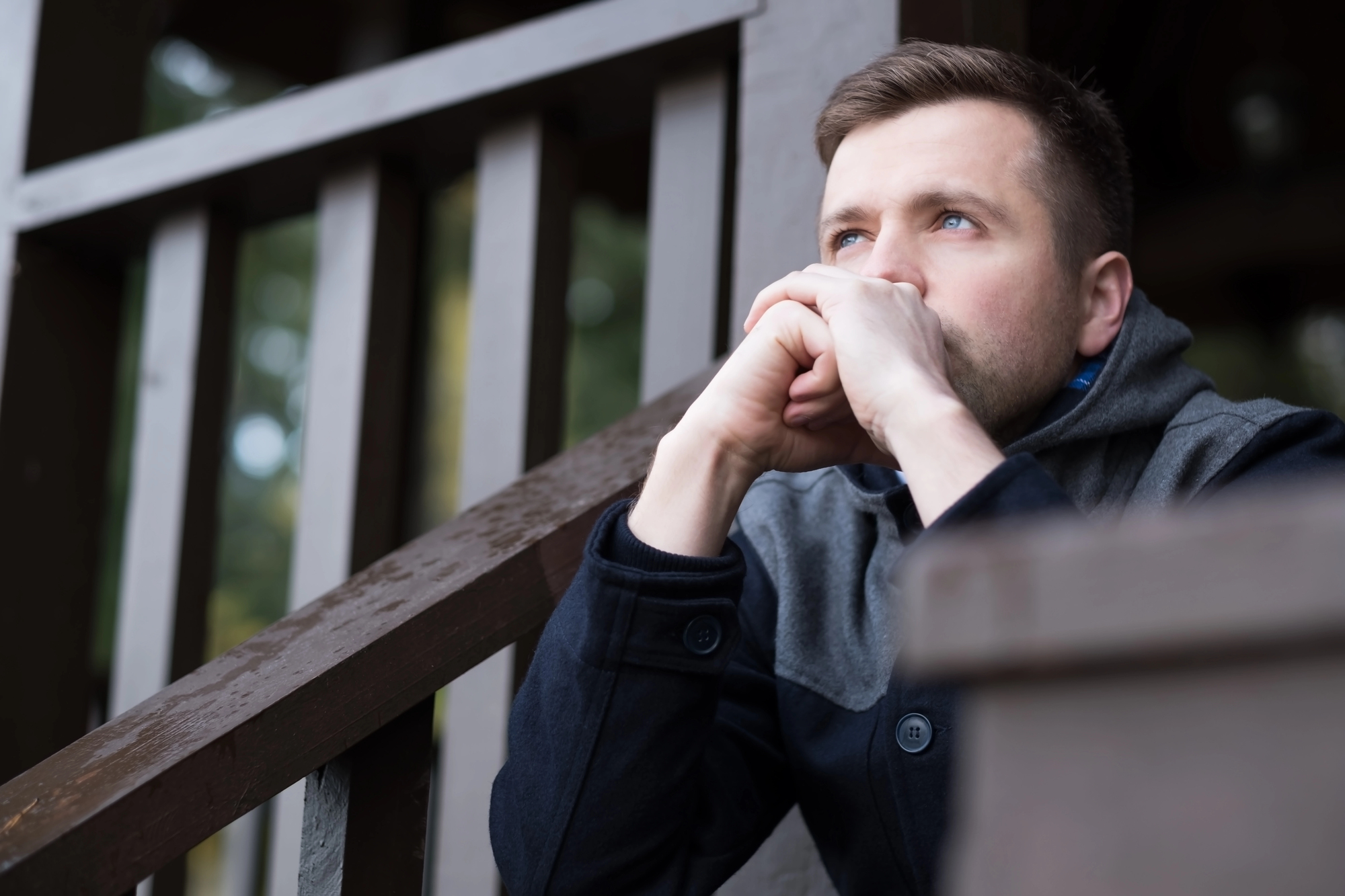 A man with short brown hair and a beard, wearing a dark jacket, sits on wooden steps with his elbows resting on his knees and his hands clasped in front of his face, appearing deep in thought. The background features wooden railings and a blurred natural setting.