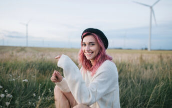 A young person with pink hair and a black beret is sitting in a grassy field near wind turbines, smiling at the camera. They are wearing a light-colored sweater and holding a small plant. The sky is pale and clear in the background.