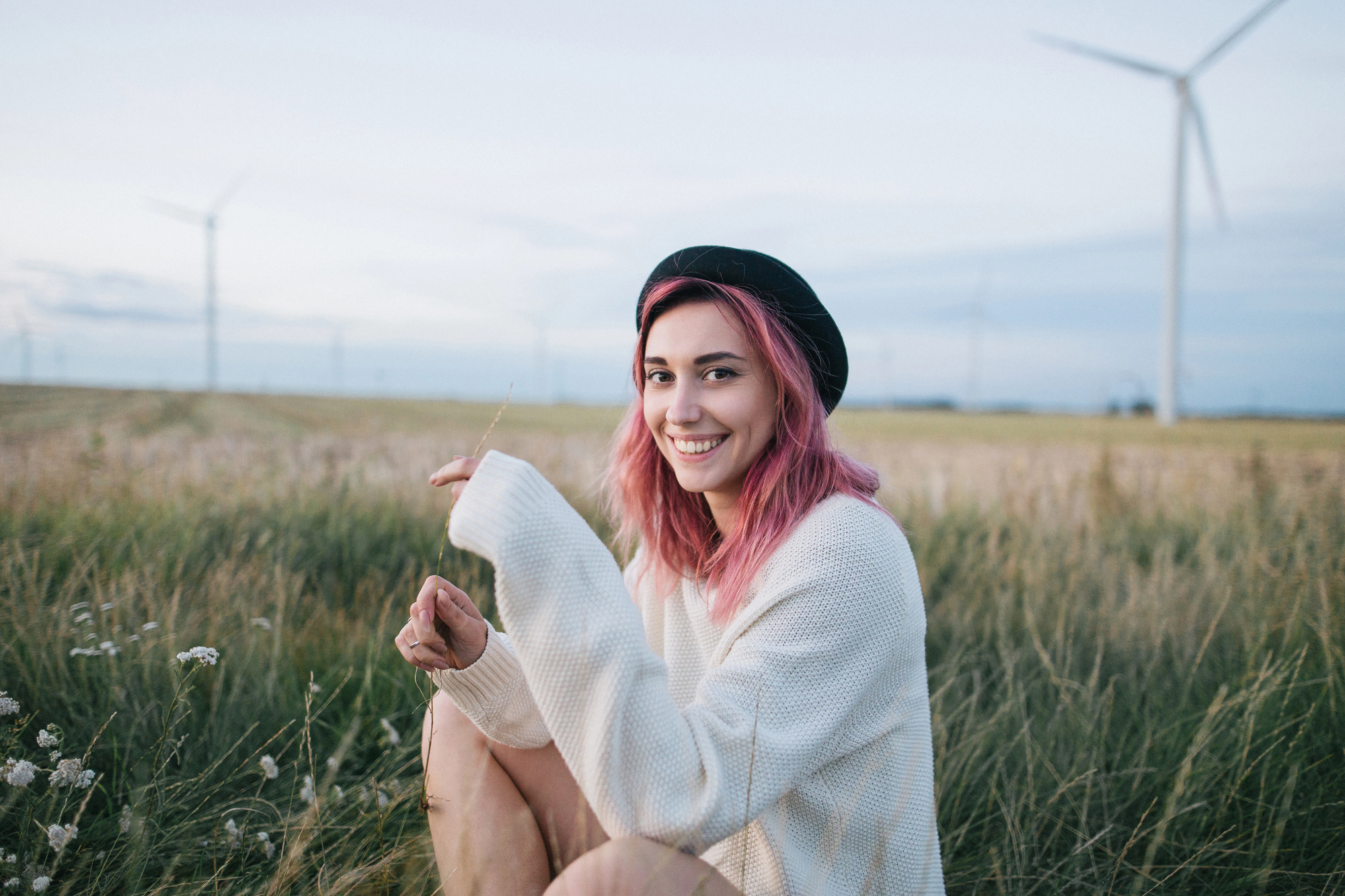 A young person with pink hair and a black beret is sitting in a grassy field near wind turbines, smiling at the camera. They are wearing a light-colored sweater and holding a small plant. The sky is pale and clear in the background.