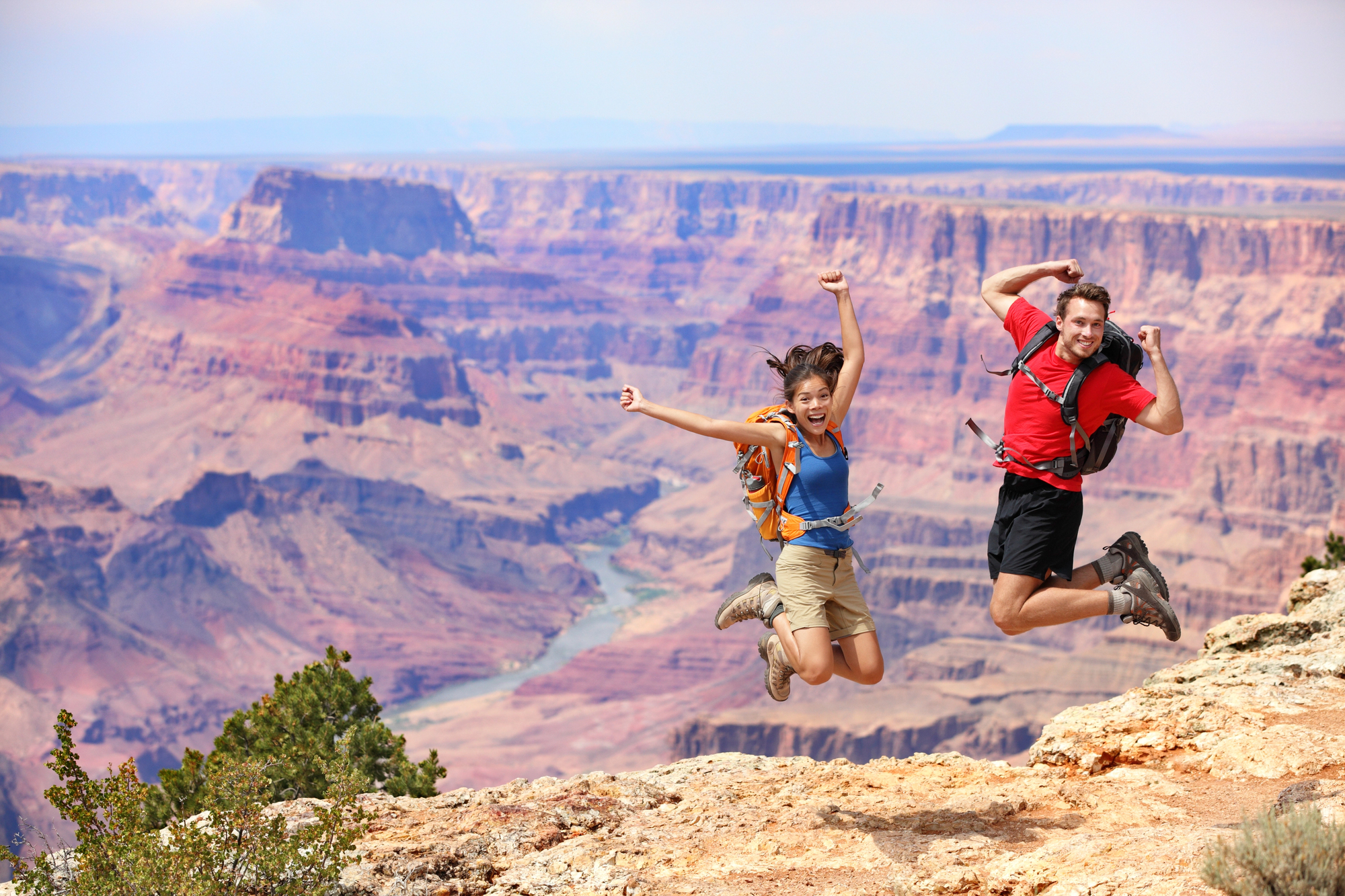 Two hikers, a man and a woman, jump joyfully at the edge of a cliff overlooking the Grand Canyon. They wear backpacks and hiking gear. The deep, colorful canyons and Colorado River form a stunning background under a clear sky.