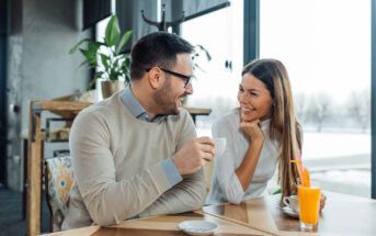 A man and a woman sit at a table in a modern, well-lit café, smiling and having a conversation. The man holds a cup of coffee, and the woman has a glass of orange juice with a straw. The background features large windows with daylight streaming in.