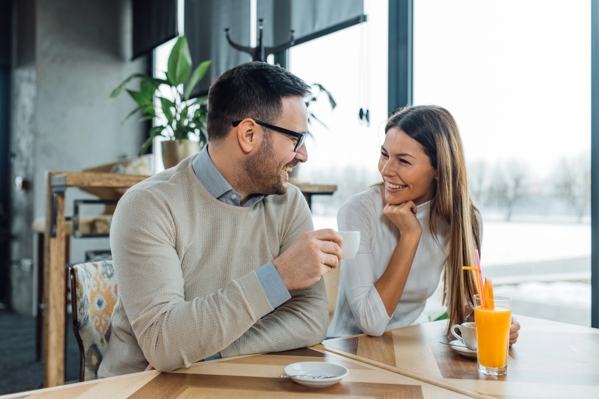 A man and a woman sit at a table in a modern, well-lit café, smiling and having a conversation. The man holds a cup of coffee, and the woman has a glass of orange juice with a straw. The background features large windows with daylight streaming in.