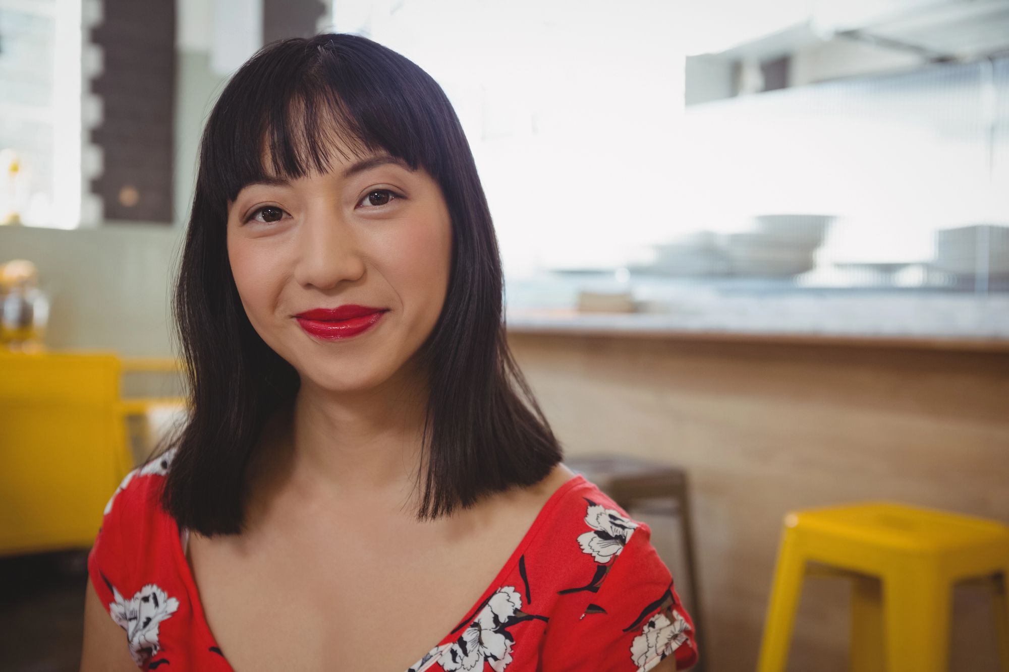 A woman with straight black hair and bangs, wearing a red floral dress, smiles at the camera. She is seated indoors, with a blurred background of a kitchen and yellow chairs.