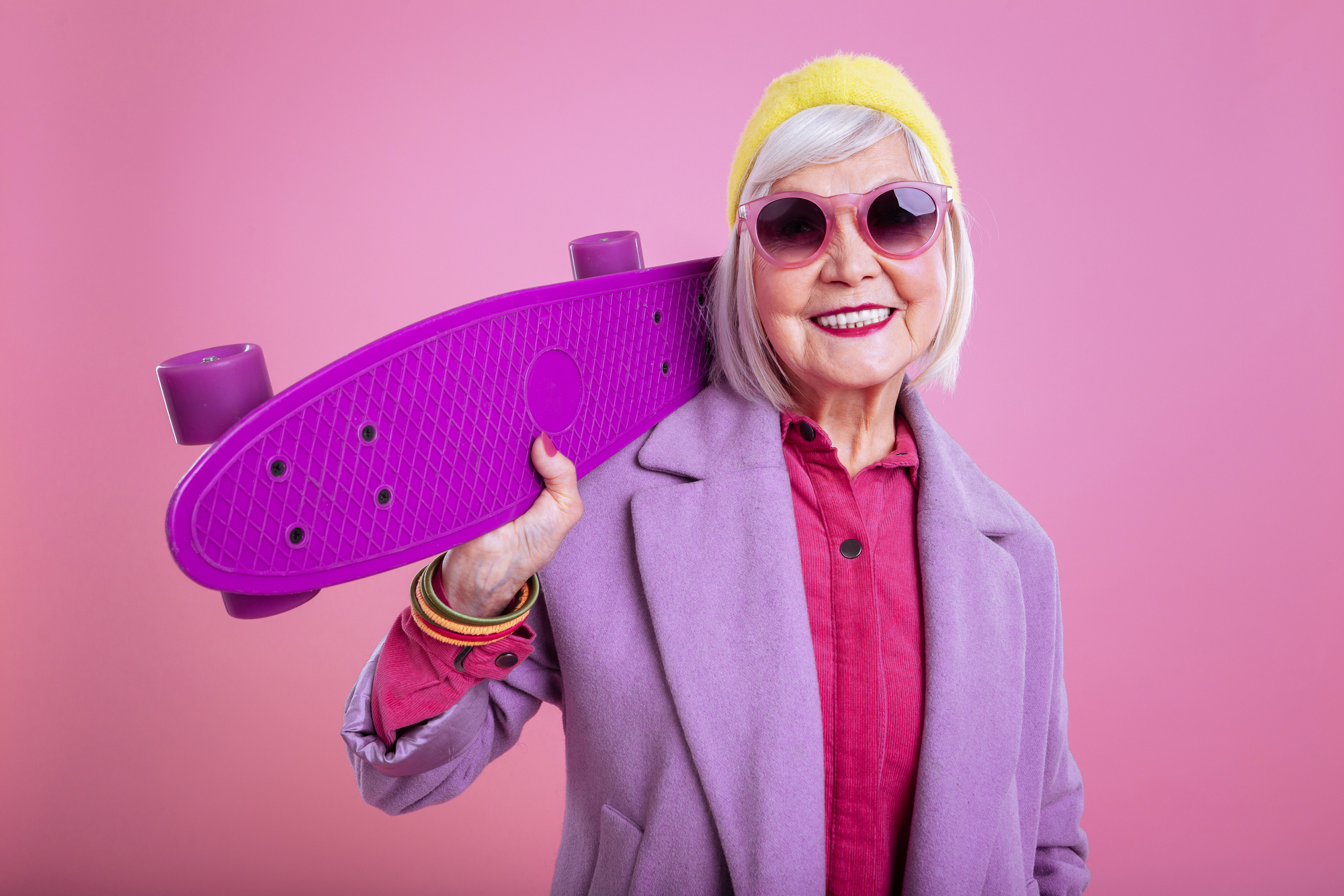 A stylish elderly woman wearing a yellow headband, pink sunglasses, a purple coat, and a pink shirt smiles while holding a bright purple skateboard over her shoulder. The background is a solid pink color, adding to the vibrant and cheerful scene.