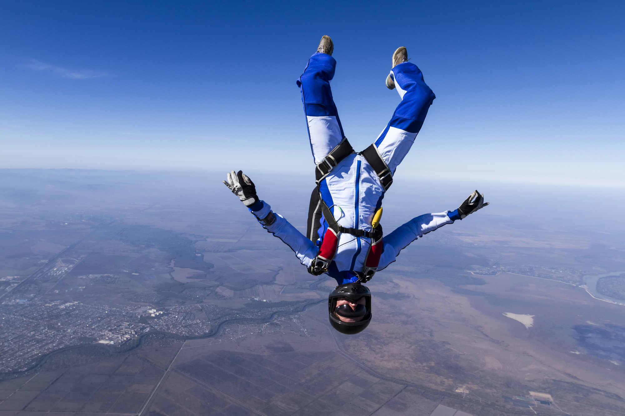 A skydiver in a blue and white jumpsuit performs a head-down freefall maneuver against a backdrop of clear blue sky and expansive landscape below, including fields, a river, and scattered buildings. The skydiver wears a helmet and goggles, arms outstretched.